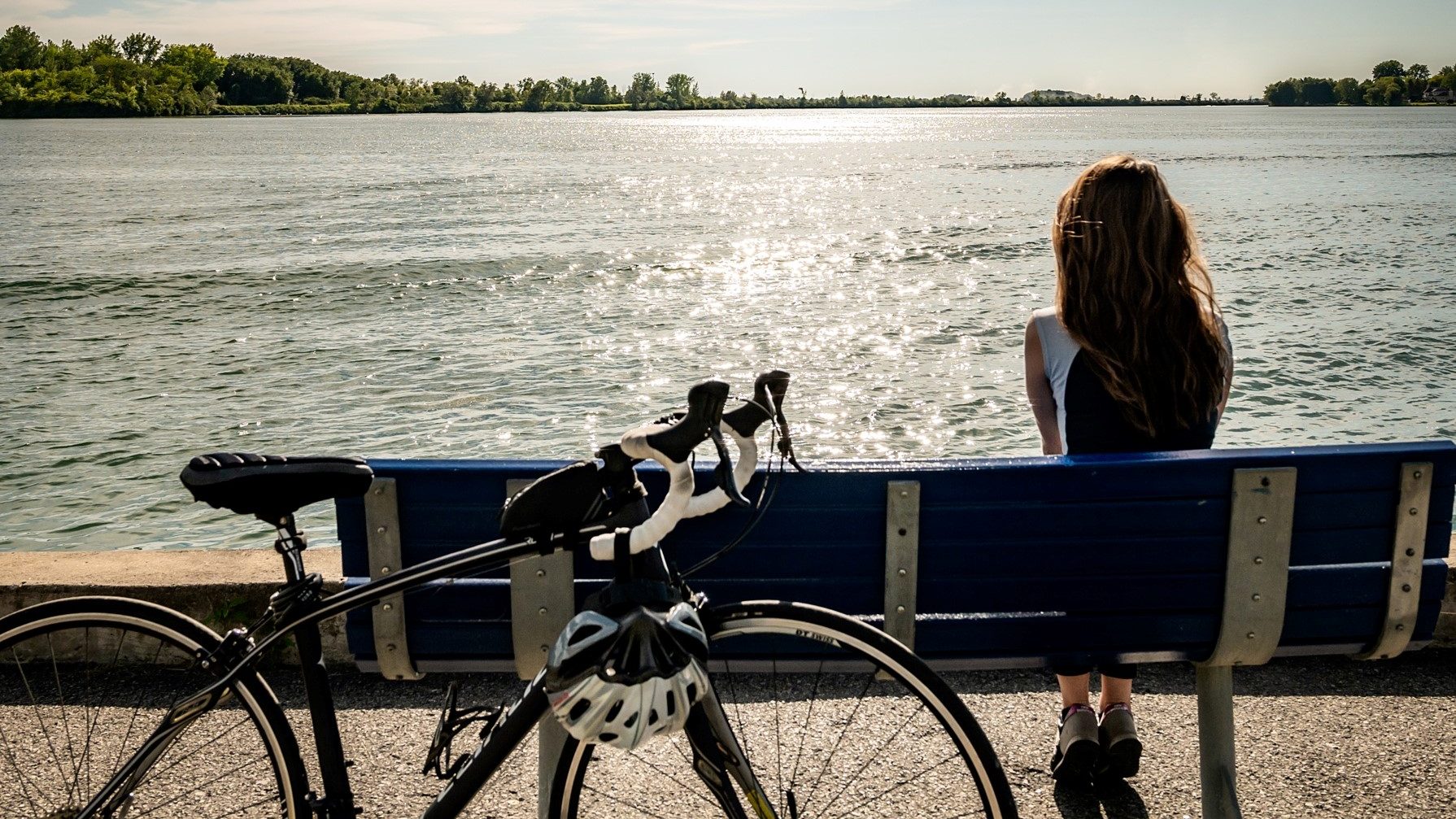 Jeune femme assise sur un banc admirant le reflet du soleil sur l'eau, son vélo appuyé contre le banc.