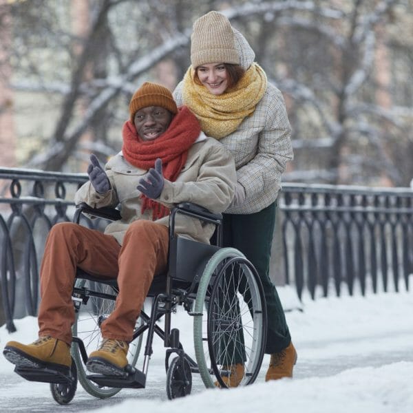 homme en fauteuil roulant avec une accompagnatrice, à l'extérieur en hiver.